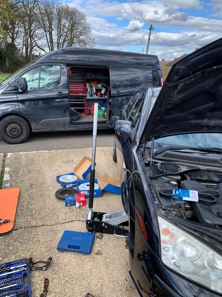 A mobile mechanic van with its tool racking open, parked beside a car mid brake job, with new discs and pads on the driveway
